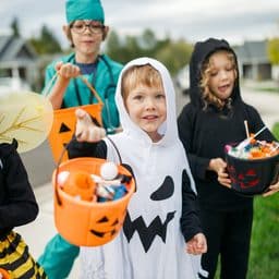 A cute group of young elementary age kids trick or treating for the Halloween holiday in their neighborhood. They wear assorted traditional costumes, carrying Jack-o'-lantern buckets filled with candy, treats, and toys.