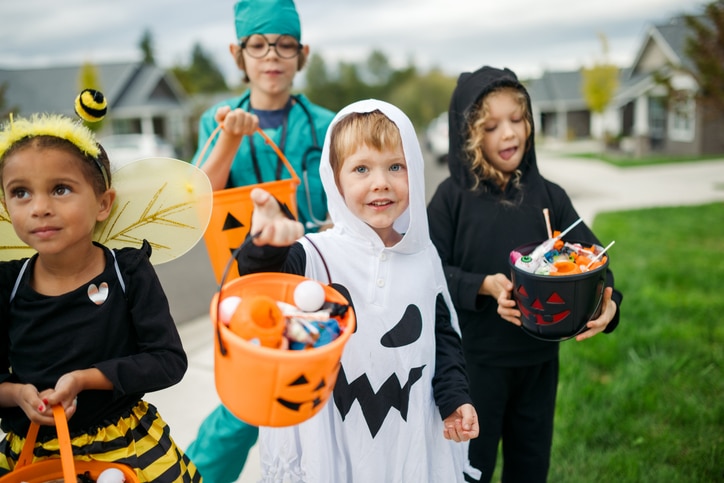 A cute group of young elementary age kids trick or treating for the Halloween holiday in their neighborhood. They wear assorted traditional costumes, carrying Jack-o'-lantern buckets filled with candy, treats, and toys.